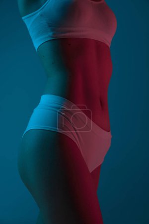 Foto de Sportive lifestyle. Cropped image of slim fit female body in underwear over blue studio background in neon light Concept of body and skin care, fitness, natural beauty, health, wellness. - Imagen libre de derechos