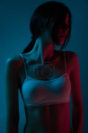 Téléchargez les photos : Portrait de belle jeune fille brune avec un corps mince posant en sous-vêtements sur fond bleu studio en lumière au néon. Concept de soins corporels et cutanés, fitness, beauté naturelle, santé, bien-être. - en image libre de droit