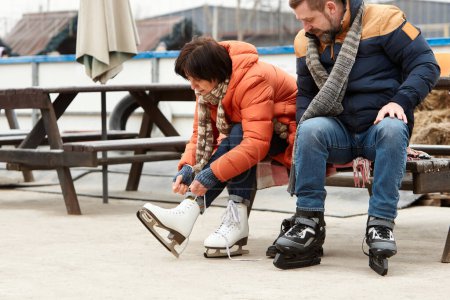 Photo for Mature man and woman getting ready to skate on ice rink outdoors. Woman tying laces on white skates. Active weekends. Concept of leisure activity, winter hobby, vacation, fun, relationship, emotions. - Royalty Free Image