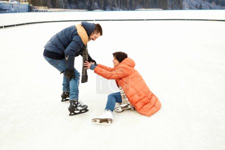 Photo for Middle-aged couple skating on open air ice rink. Man helping fallen woman to get up. Concept of leisure activity, winter hobby and sport, vacation, fun, relationship, emotions. - Royalty Free Image