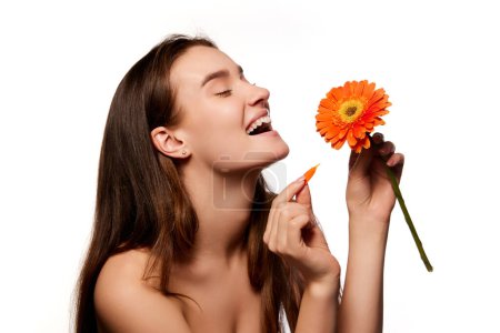Téléchargez les photos : Positivity, energy and self-care. Beautiful young girl with well-kept clean skin posing with gerbera flower over white studio background. Concept of natural beauty, youth, health, wellness, femininity - en image libre de droit
