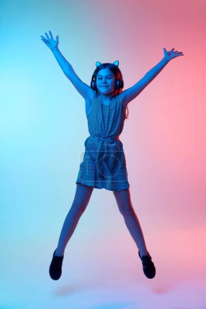 Photo for Happiness. Little beautiful girl, child in headphones jumping over gradient blue pink studio background in neon light. Concept of childhood, emotions, fun, fashion, lifestyle, facial expression - Royalty Free Image