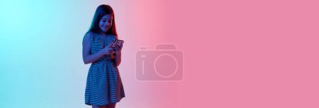 Photo for Online communication. Little smiling girl, child chatting on phone over gradient blue pink studio background in neon light. Concept of childhood, emotions, fun, fashion, lifestyle. Banner, flyer - Royalty Free Image