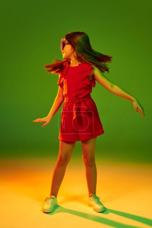 Foto de Little girl, child in red clothes posing, dancing over gradient green yellow studio background in neon light. Concept of childhood, emotions, fun, fashion, lifestyle, facial expression - Imagen libre de derechos