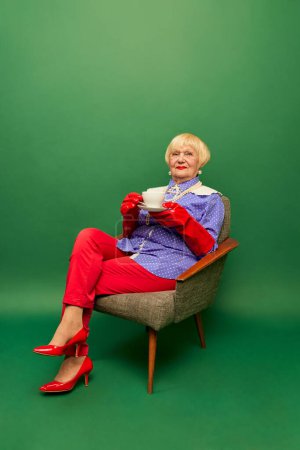 Photo for Beautiful old woman, grandmother in colorful clothes sitting on armchair, drinking coffee over green studio background. Concept of age, fashion, lifestyle, emotions, facial expression - Royalty Free Image
