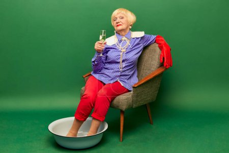 Foto de Beautiful old woman, grandmother in stylish colorful clothes sitting with legs into bowl and drinking champagne over green studio background. Concept of age, fashion, lifestyle, emotions, celebration - Imagen libre de derechos