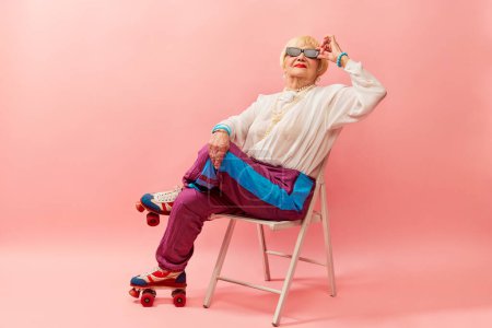 Foto de Un ambiente relajado. Hermosa anciana, abuela en pantalones deportivos elegantes posando en rodillos vintage sobre fondo de estudio rosa. Concepto de edad, moda, estilo de vida, emociones, expresión facial - Imagen libre de derechos