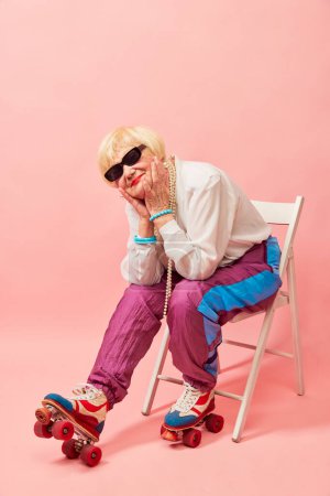 Foto de Mirada de ensueño. Hermosa anciana, abuela en pantalones deportivos elegantes posando en rodillos vintage sobre fondo de estudio rosa. Concepto de edad, moda, estilo de vida, emociones, expresión facial - Imagen libre de derechos