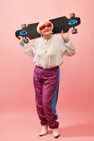 Foto de Energetic lady. Beautiful old woman, grandmother in stylish clothes posing with skateboard over pink studio background. Concept of age, fashion, lifestyle, emotions, facial expression - Imagen libre de derechos