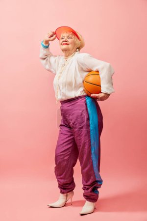 Photo for Sportive past. Beautiful old woman, grandmother in stylish clothes posing with basketball ball over pink studio background. Concept of age, fashion, lifestyle, emotions, facial expression - Royalty Free Image