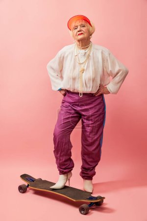 Schöne alte Frau, Großmutter in stylischer Kleidung, die mit Skateboard vor rosa Studiohintergrund posiert. Konzept von Alter, Mode, Lebensstil, Emotionen, Mimik