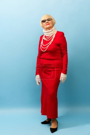 Fashionable look. Beautiful old woman, grandmother in stylish red dress and pearl necklace posing over blue studio background. Concept of age, fashion, lifestyle, emotions, facial expression-stock-photo
