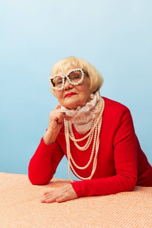 Foto de Beautiful old woman, grandmother in stylish red dress, pearl necklace and glasses posing over blue studio background. Concept of age, fashion, lifestyle, emotions, facial expression - Imagen libre de derechos