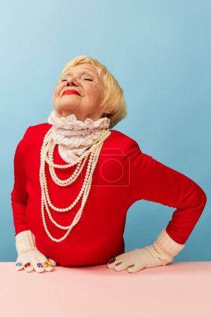 Téléchargez les photos : Délicieux. Belle vieille femme, grand-mère en robe rouge élégante et collier de perles posant sur fond bleu studio. Concept d'âge, mode, mode de vie, émotions, expression faciale - en image libre de droit
