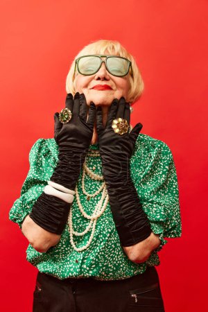 Foto de Luxurious accessories. Beautiful old woman, grandmother in stylish clothes, gloves and glasses posing over red studio background. Concept of age, fashion, lifestyle, emotions, facial expression - Imagen libre de derechos
