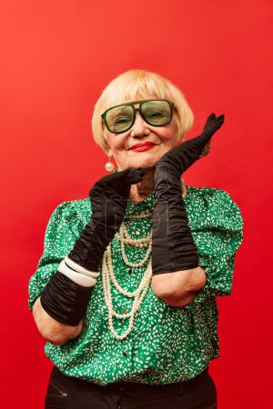 Foto de Extravagant look. Beautiful old woman, grandmother in stylish clothes, gloves and glasses posing over red studio background. Concept of age, fashion, lifestyle, emotions, facial expression - Imagen libre de derechos