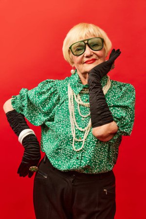 Foto de Extravagant look. Beautiful old woman, grandmother in stylish clothes, gloves and glasses posing over red studio background. Concept of age, fashion, lifestyle, emotions, facial expression - Imagen libre de derechos