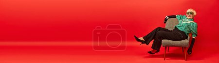 Photo for Relaxed. Beautiful old woman, grandmother in stylish clothes posing, sitting on armchair over red studio background. Age, fashion, lifestyle, emotions, facial expression. Banner. Copy space for ad - Royalty Free Image