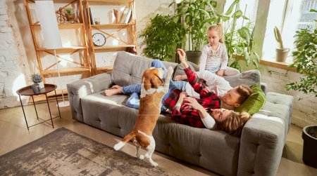 Foto de Lovely cute family, parents and child relaxing at home, playing with beagle dog in their living room on sunny day. Weekends. Concept of relationship, family, parenthood, childhood, animal life - Imagen libre de derechos