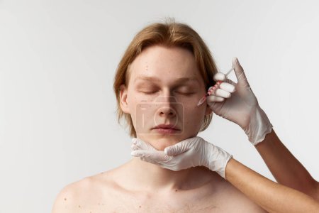 Foto de Making botox in cheeks. Young redhead man having cosmetological injections. Male model posing against grey background. Mens health, cosmetology, plastic surgery, body and skin care, medicine concept - Imagen libre de derechos