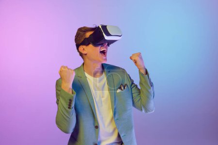Téléchargez les photos : Futurism, virtual reality. Young handsome man in stylish jacket posing in VR glasses over purple background in neon light. Concept of emotions, lifestyle, youth, modern fashion. Ad - en image libre de droit