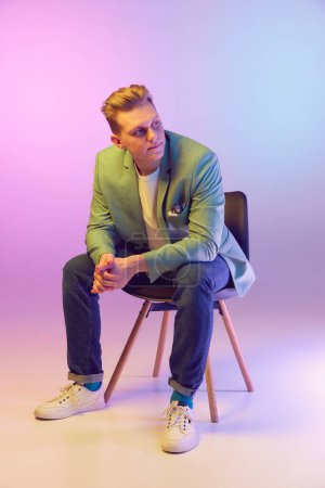 Photo for Elegant look. Young handsome man in stylish jacket posing, sitting on chair over gradient purple background in neon light. Concept of emotions, lifestyle, youth, modern fashion. Ad - Royalty Free Image