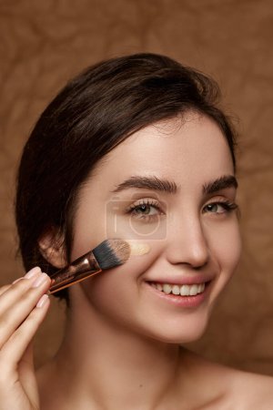 Téléchargez les photos : Beautiful young smiling brunette girl applying face foundation with brush, doing nude makeup over vintage brown studio background. Concept of natural beauty, skincare, cosmetology, cosmetics, health - en image libre de droit