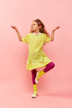 Téléchargez les photos : Balancing on one leg. Little cute girl, child with curly hair, colorful hair clips posing in yellow dress over pink studio background. Childhood, emotions, fun, fashion, lifestyle, facial expression - en image libre de droit