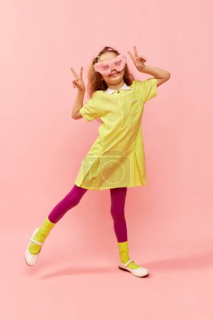 Téléchargez les photos : Bonheur et positivité. Petite fille mignonne, enfant aux cheveux bouclés posant en robe jaune et lunettes de fourrure rose sur fond de studio rose. Enfance, émotions, amusement, mode, style de vie, expression faciale - en image libre de droit