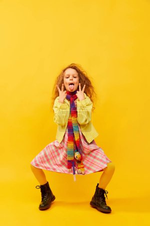 Téléchargez les photos : Peace symbol. Little cute girl, child with curly hair posing in bright clothes with tongue out over yellow studio background. Concept of childhood, emotions, fun, fashion, lifestyle, facial expression - en image libre de droit