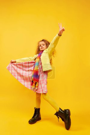 Téléchargez les photos : Little cute girl, child with curly hair posing in bright clothes, scarf and boots over yellow studio background. Concept of childhood, emotions, fun, fashion, lifestyle, facial expression - en image libre de droit