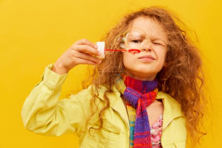Photo for Positivity. Little cute girl, child with curly hair posing in bright clothes, blowing bubbles over yellow studio background. Concept of childhood, emotions, fun, fashion, lifestyle, facial expression - Royalty Free Image