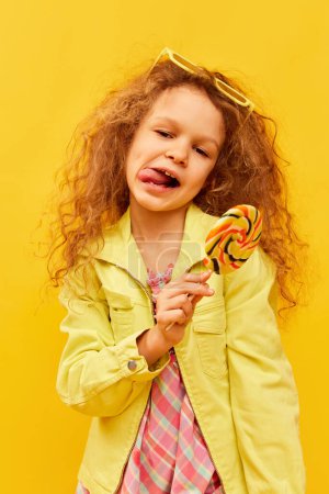 Photo for Positive mood. Little cute girl, child with curly hair posing in bright clothes with lollipop over yellow studio background. Concept of childhood, emotions, fun, fashion, lifestyle, facial expression - Royalty Free Image