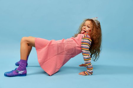 Téléchargez les photos : Playful kid having fun. Little cute girl, child with curly hair posing in pink dress over blue studio background. Concept of childhood, emotions, fun, fashion, lifestyle, facial expression - en image libre de droit