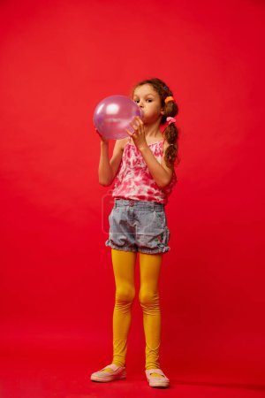 Téléchargez les photos : Little cute girl, child with braids emotionally posing in yellow tights, blowing air balloon on bright red studio background. Concept of childhood, emotions, fun, fashion, lifestyle, facial expression - en image libre de droit