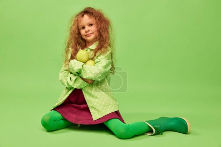 Photo for Vitamins and taste. Little cute girl, child with curly hair posing with apples over green studio background. Concept of childhood, emotions, fun, fashion, lifestyle, facial expression - Royalty Free Image
