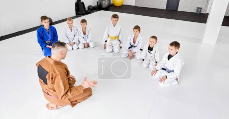 Foto de Grupo de niños pequeños, niños en kimono sentados en el suelo alrededor del entrenador. Niños entrenando en interiores. Judo, entrenamiento jiu-jitsu. Concepto de artes marciales, deporte de combate, educación deportiva, infancia, - Imagen libre de derechos