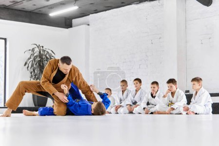 Photo for Teacher, professional judo, jiu jitsu coach training, teaching kinds, boys to fight. Children learning indoors. Concept of martial arts, combat sport, sport education, childhood - Royalty Free Image