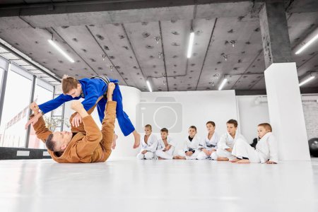 Téléchargez les photos : Professional judo, jiu-jitsu coach showing exercise, training indoors. Children attentively looking at trainer. Concept of martial arts, combat sport, sport education, childhood, hobby - en image libre de droit