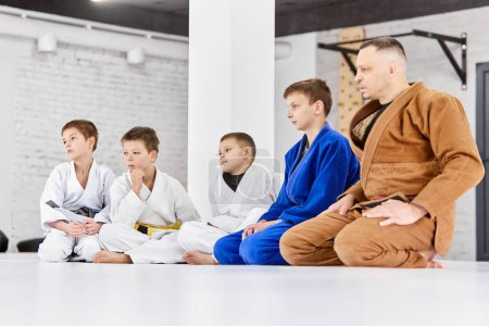 Photo for Attentive look. Coach of judo, jiu-jitsu sitting with little boys, children in white kimono, watching exercise technique. Concept of martial arts, combat sport, sport education, childhood, hobby - Royalty Free Image