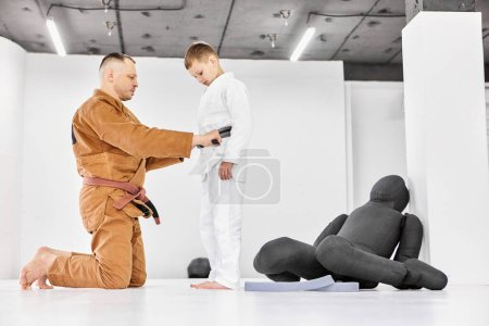 Téléchargez les photos : Homme, judo, entraîneur de jiu-jitsu aidant garçon, enfant à attacher la ceinture sur le kimono. Activité d'entraînement, apprendre à se battre. Concept d'arts martiaux, sport de combat, éducation sportive, enfance, passe-temps, mode de vie - en image libre de droit