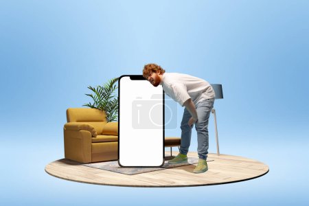 Foto de Young man leaning on giant 3D model of mobile phone with empty screen for ad, text over blue background with home interior. Online communication, shopping. Mockup for design, logo. - Imagen libre de derechos