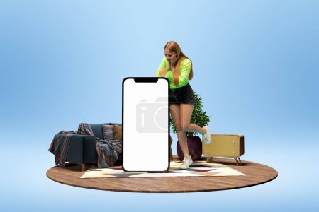 Téléchargez les photos : Excited young girl emotionally looking on giant 3D model of mobile phone with empty screen for text, ad over blue background with home interior. Betting, online shopping. Mockup for design, logo. - en image libre de droit