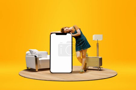 Foto de Positive smiling young girl leaning on huge 3D model of mobile phone with empty screen for text, ad over yellow background with home interior. Freelance. Mockup for ad, design, logo. - Imagen libre de derechos