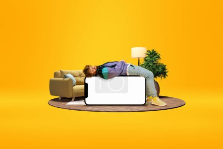 Téléchargez les photos : Relaxed young man lying on huge 3D model of mobile phone with empty screen for text, ad over bright yellow background with home interior. Online shopping, betting. Mockup for ad, design, logo. - en image libre de droit