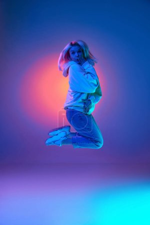 Foto de Surprise. Emotive young woman in casual clothes jumping over gradient multicolored studio background in blue neon light. Concept of emotions, facial expression, lifestyle, inspiration, sales, ad - Imagen libre de derechos