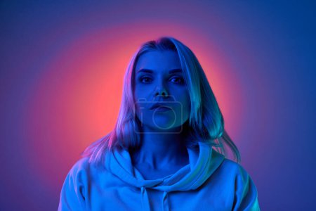 Foto de Portrait of young beautiful blonde woman attentively looking at camera over gradient studio background in blue neon light. Concept of emotions, facial expression, lifestyle, inspiration, sales, ad - Imagen libre de derechos