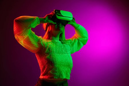 Téléchargez les photos : Modern gadgets and technologies. Young woman in VR glasses over magenta studio background in green neon light. Concept of emotions, facial expression, lifestyle, inspiration, sales, ad - en image libre de droit