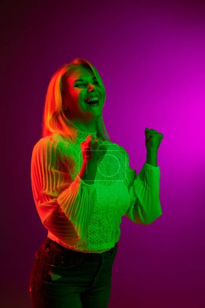 Photo for Success. Happy excited young woman posing with winning look over magenta studio background in green neon light. Concept of emotions, facial expression, lifestyle, inspiration, sales, ad - Royalty Free Image