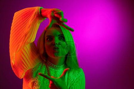 Foto de Taking photos gesture. Young pretty woman posing over magenta studio background in green neon light. Concept of emotions, facial expression, lifestyle, inspiration, sales, ad - Imagen libre de derechos